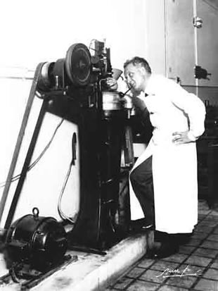 Szent-Györgyi Albert álló, egészalakos képe. A laboratóriumi paprikaőrlő mellett áll, vizsgálódik.